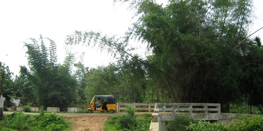 A yellow auto-rickshaw parked on a dirt road by a concrete bridge, surrounded by greenery