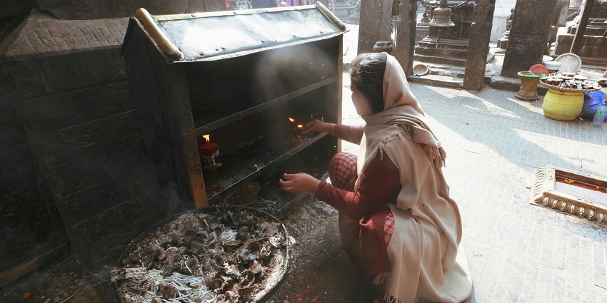 A woman lighting a candle in front of the stand in a temple with a bell in the backdrop.
