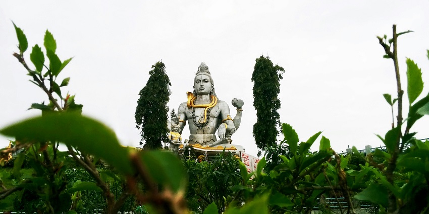 A statue of Shiva with four arms, adorned with gold, viewed through green foliage