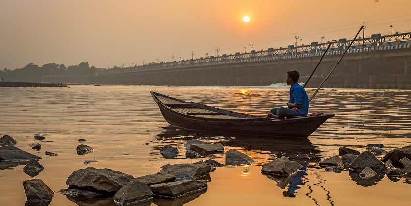 An oarsman sits on his boat at sunset on river Damodar on a foggy winter afternoon.