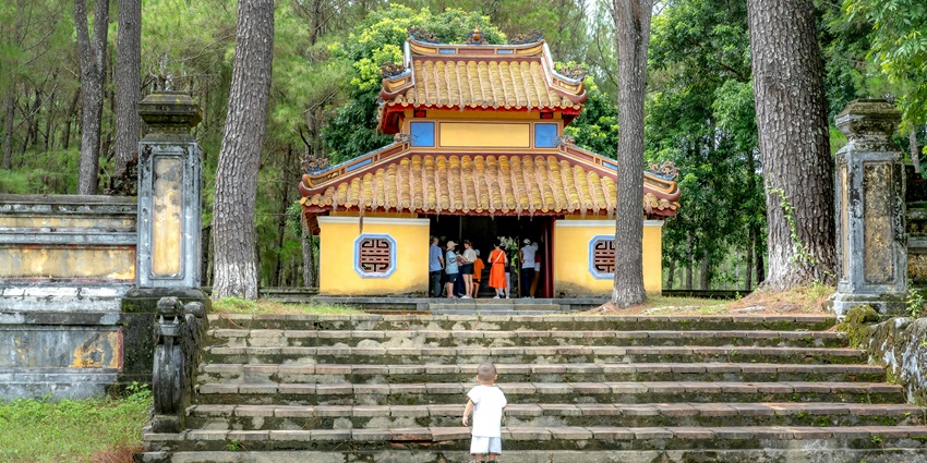 A child stands in front of an ancient temple, partially surrounded by lush green foliage.