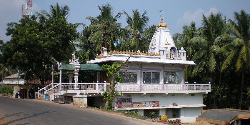 A distant view of a white-coloured temple is showcased located on the side of a road.