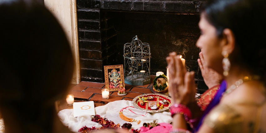 Two women praying in front of a photograph of a deity by lighting lamps and candles.