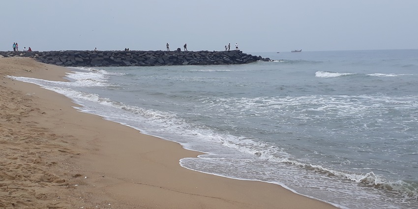 Waves hitting a sandy shore near a rocky jetty with people walking and standing on it.
