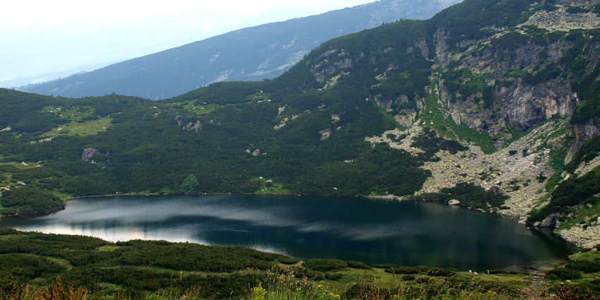One of the seven lakes of Rila is surrounded by greenery, cliffs and dark mountains.