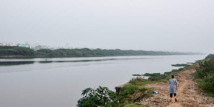 A person walking on a path beside a calm lake with greenery and distant buildings