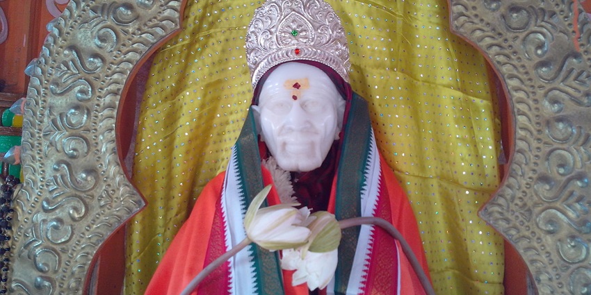 Statue of Sai Baba against a golden backdrop, decorated with flowers and colourful garments.