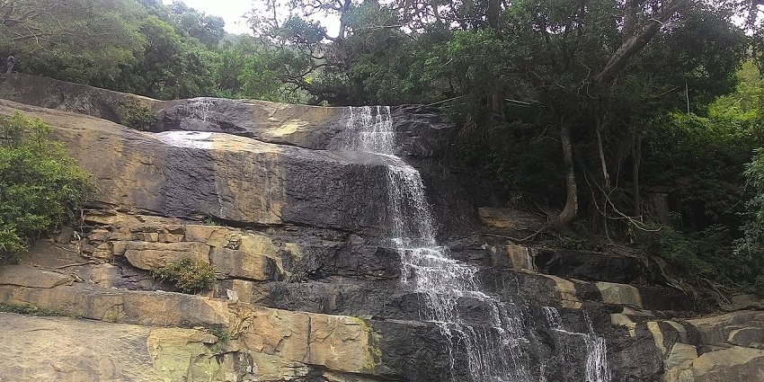 Suruli Falls cascading down rocky cliffs, surrounded by greenery and people standing below