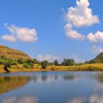 A peaceful pond near Tringalwadi Fort surrounded by rolling hills and a clear blue sky.