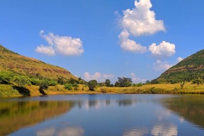 A peaceful pond near Tringalwadi Fort surrounded by rolling hills and a clear blue sky.