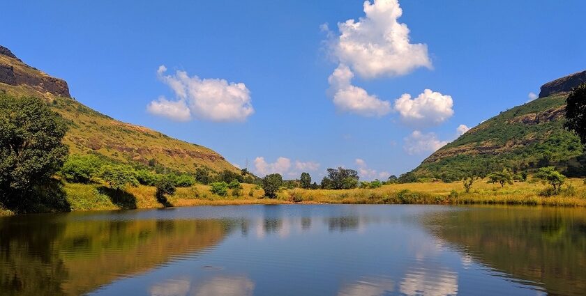 A peaceful pond near Tringalwadi Fort surrounded by rolling hills and a clear blue sky.