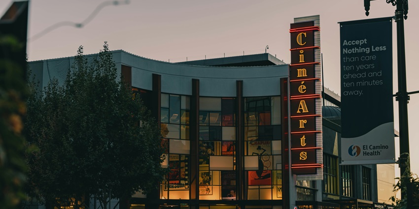 A movie theater lit up at dusk, with posters in the windows and a banner nearby.