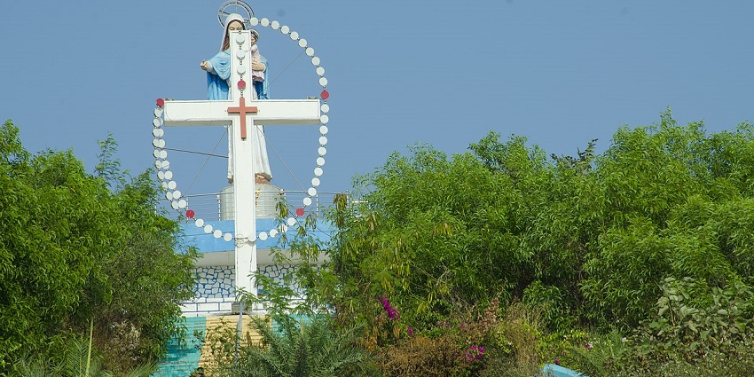 Statue of the Virgin Mary with a white cross near Velankanni Beach, surrounded by greenery.