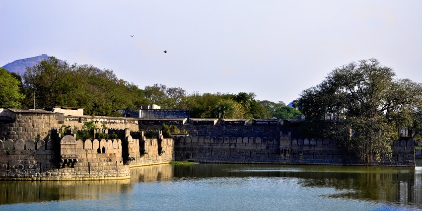 Structure of Vellore Fort is surrounded by greenery and birds flying near the water.