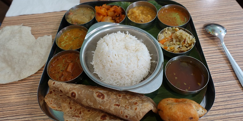 A South Indian thali with rice, curries, dal, sambar, and sides on a round stainless steel platter.