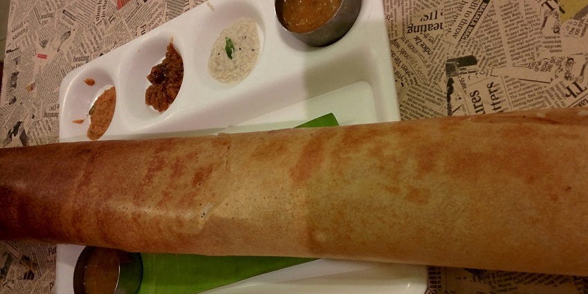 A crispy thin golden brown paper Dosa served with chutneys and sambar on a white plate.