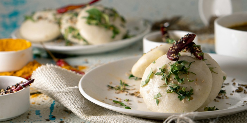 Idli is served with herbs and topped with chillis and other plates on the table with tissues.
