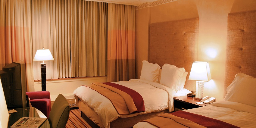 An illuminated hotel room featuring two double beds, a lamp, a glass table and a chair.