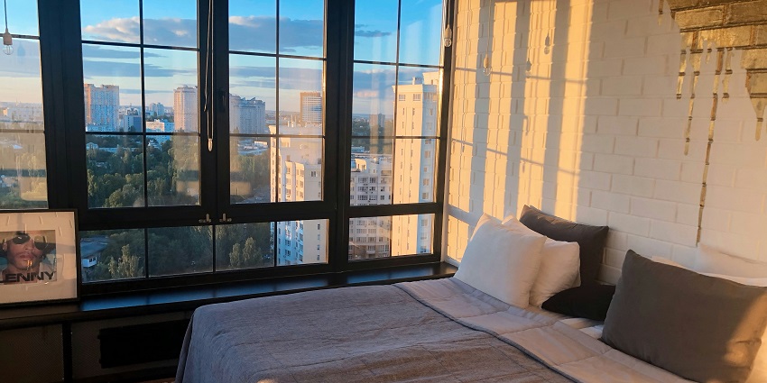 A bed in a hotel room with large glass windows displaying a magnificent view of the city