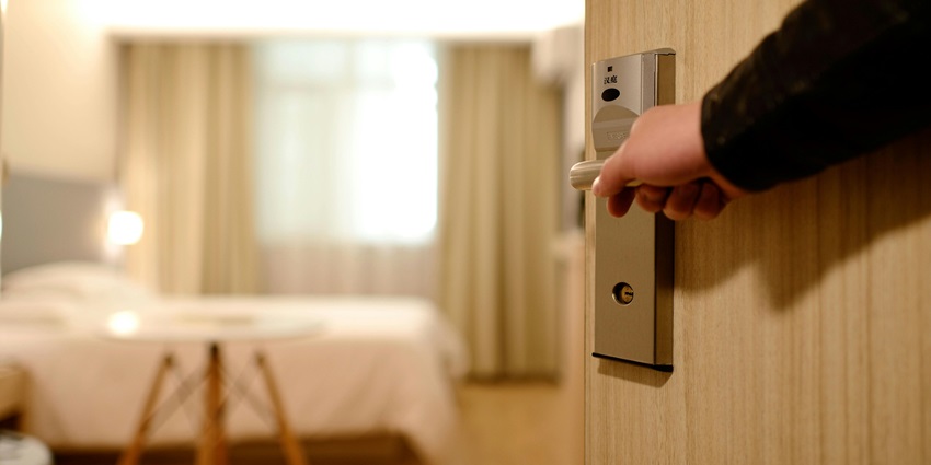 A person holding the lever of a door for an entry inside the hotel room having a bed and table.