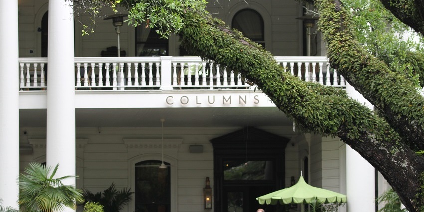 Exterior of the Columns Hotel with white columns, balcony railings, and people coming out.