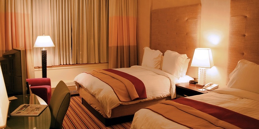 The hotel room is warmly lit with two made beds, pillows, lamps, a desk and closed curtains
