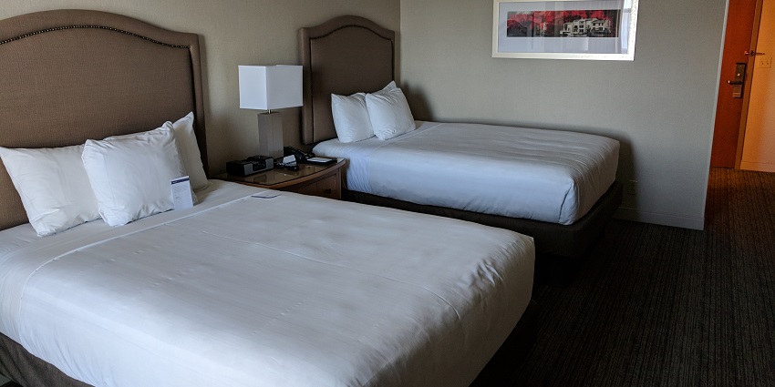 A hotel room with two made beds with cushions, a lamp and a painting hanging on the wall.