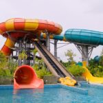 A colourful water slide stands tall, surrounded by a bright blue pool and a sunny sky.