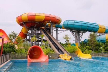 A colourful water slide stands tall, surrounded by a bright blue pool and a sunny sky.