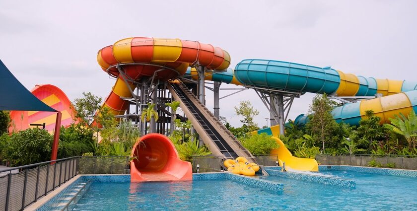 A colourful water slide stands tall, surrounded by a bright blue pool and a sunny sky.