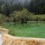 An image of a Waterfall in Alaska cascading down a mountain, surrounded by lush green trees and rocks