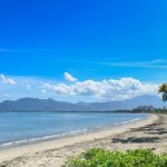 A view of a public beach with clear blue waters, soft sand, and palm trees nearby.