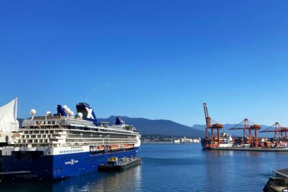 A view of a ship docked in a harbour surrounded by lush greenery, city view, and mountains.