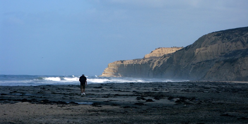 A man is walking alone on Black’s Beach, San Diego, towards the high-rise cliffs