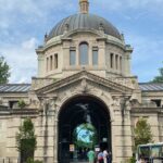 Bronx Zoo entrance on Fordham Road shows visitors walking near trees and park signage.