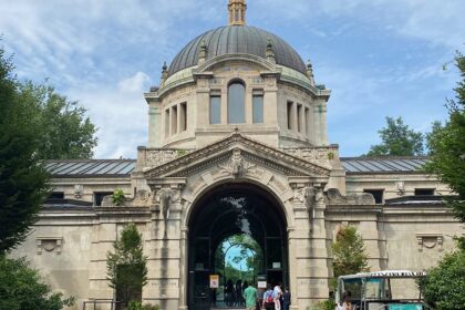 Bronx Zoo entrance on Fordham Road shows visitors walking near trees and park signage.