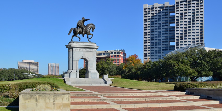 Sam Houston's monument suris rounded by greenery in Hermann Park and tall buildings.