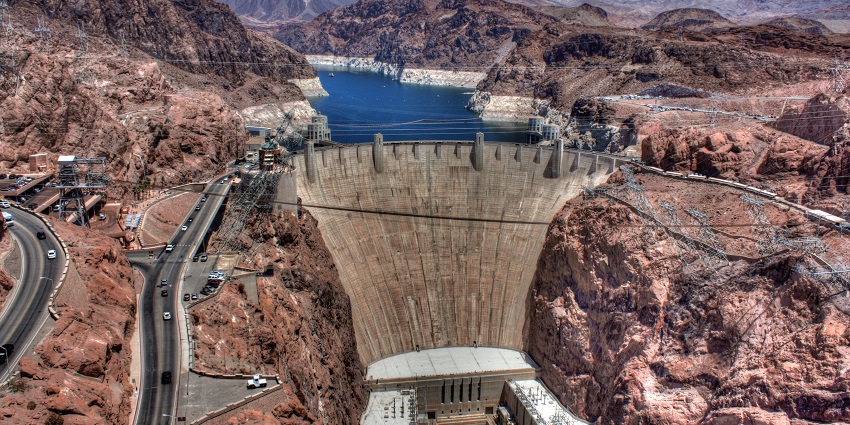 Hoover Dam from the top, surrounded by rocky hills and roads with vehicles and a river