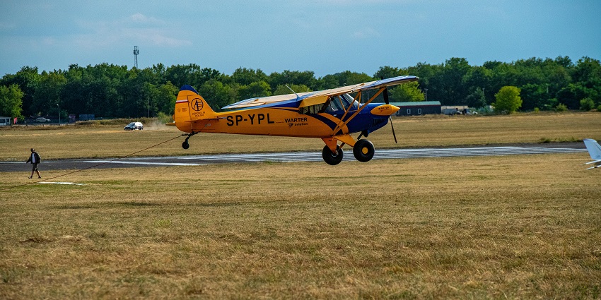 A small aeroplane taking off from the grass airfield with a man walking and a distant car