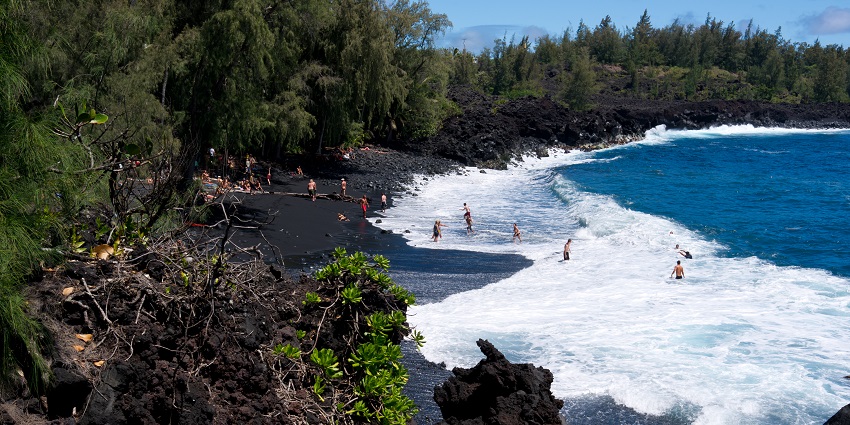 Visitors are enjoying the waves on a small strip of black sand amidst green surroundings