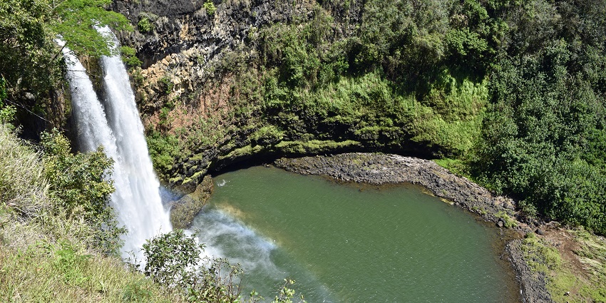 An image of a waterfall flowing through rocks, near a grassy field under a cloudy sky.