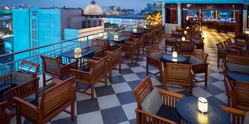 A rooftop bar with chairs and lamps on each table and the city buildings visible