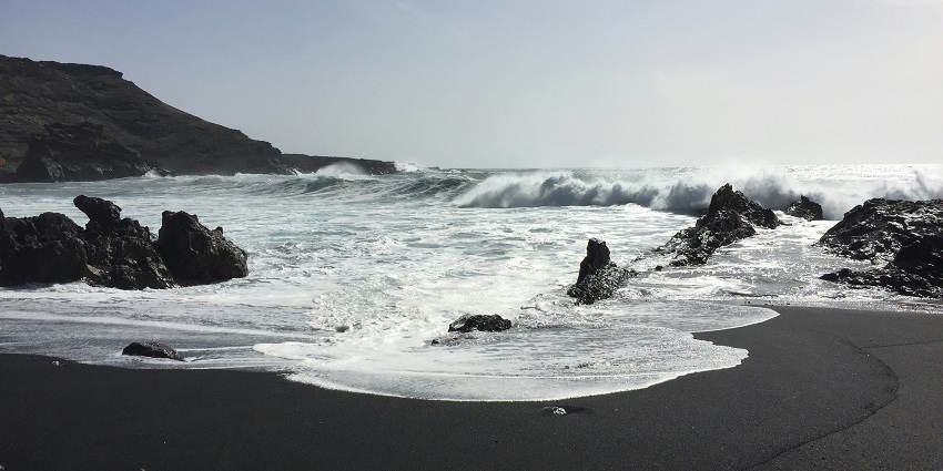 Waves are coming to the black, wet sand on a volcanic beach surrounded by rocky remains
