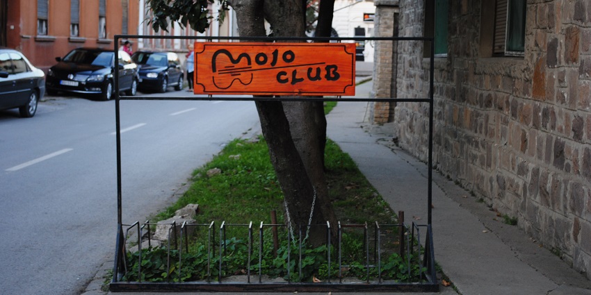 Mojo Club written on an Orange signboard on the sidewalk of a street with cars parked.