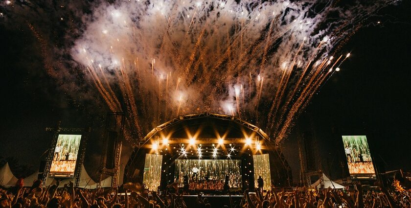 Concert at a music festival at night with fireworks and a crowd raising their hands.