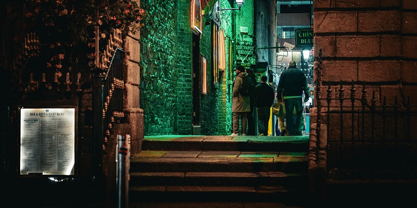 Bar entrance with a signboard on the entrance, green lighting, stairs and people visiting