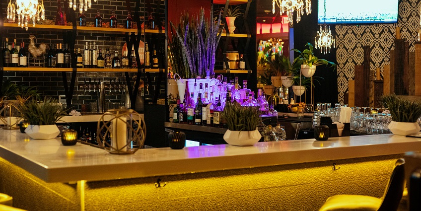 A luxury bar counter with warm lighting, alcohol bottles on the shelves and a television.