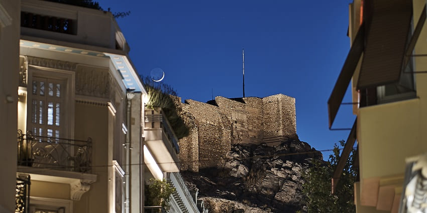 Neighbourhood of Plaka at night with buildings, green trees and a structure on a cliff