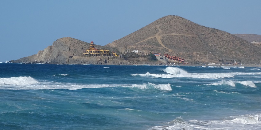 A beach with gentle waves, surrounded by mountains and green plants under a clear sky.