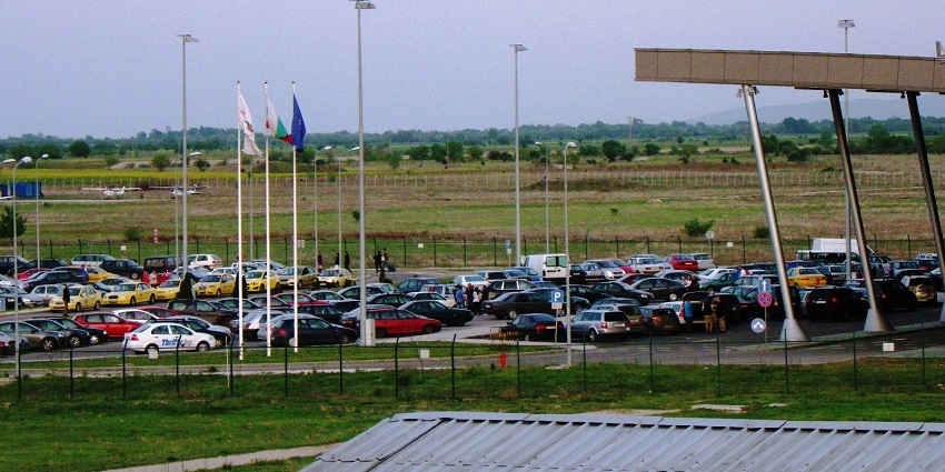 Several cars parked at the Plovdiv airport parking area, with many flagpoles standing